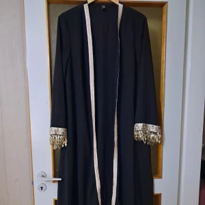 Svart lång rock med guldpaljetter - Lång svart rock med lyxiga detaljer i guld och silver längs kanterna och ärmsluten. Ärmarna är dekorerade med glittrande paljetter och fransar som ger en unik och festlig vibe. Perfekt för dig som vill sticka ut med en elegant och extravagant look. Den är ganska genomskinlig. 