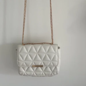 Vit quiltad axelväska med kedja - Snygg vit axelväska med quiltat triangelmönster och guldfärgad kedjerem. Väskan har ett stilrent metallspänne framtill och är perfekt för att lyfta vilken outfit som helst. Insidan har ett fack med dragkedja för småsaker. Materialet är mjukt och känns lyxigt.