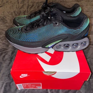 Nike Air Max DN svart/grön/blå sneakers - Nike Air Max DN sneakers i svart med blå och gröna detaljer. Ovandelen har ett futuristiskt mönster med små blå rutor och reflekterande inslag. Sulan är grå med synliga Air Max-bubblor och små Swoosh-loggor på sidorna. Skickas med shoeboxen.