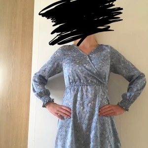 Blommig blå omlottklänning med volang - Supersöt blå kortklänning med knapp upptill som kan va uppknäppt om man önskar v-ringning, blommigt mönster, omlottliv och knyt i midjan. Långa ärmar med smock vid handlederna och volangkant nertill. Perfekt för vår och sommar när du vill känna dig extra fin💕, pris kan diskuteras❤️