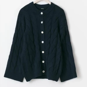 Mörkblå kabelstickad kofta med knappar - Kabelstickad kofta i mörkblått med stora vita knappar framtill. Tröjan har grovstickat mönster, ribbade muddar och rund halsringning. Perfekt för lager-på-lager och chill höst- och vinterstil.