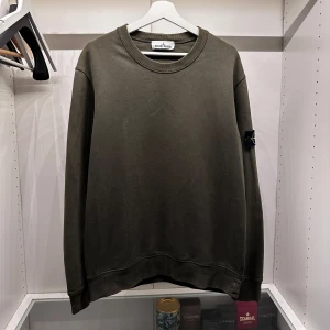 Olivgrön sweatshirt från Stone Island - Snygg olivgrön sweatshirt från Stone Island med klassisk rund hals och ikonisk patch på vänster ärm. Tröjan är i mjuk bomull och har en relaxed passform med ribbade muddar vid ärmslut och nederkant. Perfekt för dig som gillar streetwear och stilren design.