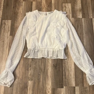 Vit volangblus från NA-KD, stl 34 - Supersöt vit blus från NA-KD med volangdetaljer framtill och längs fållen. Blusen har långärmade, transparenta ärmar med diskret prickigt mönster och elastiska muddar. Den är lätt croppad och har en feminin, luftig känsla – perfekt till jeans eller kjol. Säljer för den är för liten 💗💗