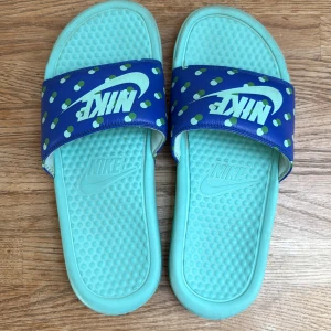 Turkosa och blå Nike sandaler - Nike sandaler i turkos med blå rem och gröna prickar. Stora vita Nike-loggor på remmen. Sval och bekväm design med platt sula och strukturmönster på fotbädden. Perfekta för sommaren eller stranden.