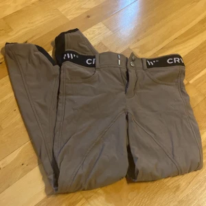 Bruna ridbyxor från CRW, stl 160 - Säljer ett par bruna ridbyxor från CRW i storlek 160. Byxorna har förstärkta partier på insidan av benen och elastisk midja med logga. Perfekta för dig som gillar ridning och vill ha bekväma och slitstarka byxor.