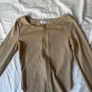 Beige ribbad kofta med dragkedja Nova - Snygg beige ribbad kofta från Nova med lång ärm och dragkedja framtill. Tröjan har en tight passform och är tillverkad i mjukt, stretchigt material som sitter skönt på kroppen. Perfekt att styla med jeans eller kjol för en clean look.