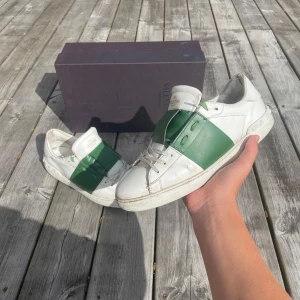 Valentino Garavani Open sneakers vit/grön - Snygga Valentino Garavani Open sneakers i vitt skinn med en bred grön läderdetalj över mitten. Klassisk rund tå, vita snören och diskret logga på plösen. Platt sula med nitar baktill ger en cool touch. Perfekta för dig som gillar statement-sneakers. Ny pris är 5500kr och kvitto finns ifall det behövs för att visa att de är äkta!