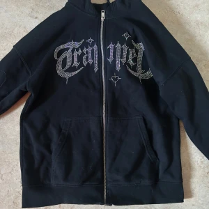 Svart zip hoodie från House - Svart hoodie från House i storlek XL med dragkedja och huva. Framsidan har en stor glittrig text i strass och coolt typsnitt. Tröjan har känguruficka och ribbade muddar. Perfekt för dig som gillar streetstyle och vill sticka ut. (AI!) Skulle säga att storleken är S-M ej XS 