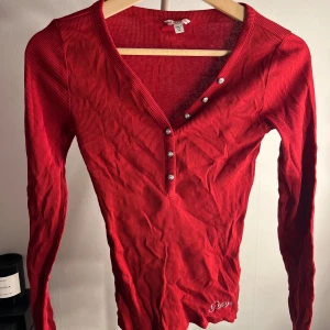 Röd ribbad långärmad tröja Guess XS - Ribbad röd långärmad tröja från Guess i storlek XS. Tröjan har v-ringning med silvriga knappar och glittrig Guess-logga i strass vid nederkanten. Snygg slim fit och stretchigt material som sitter tajt och markerar figuren. Bara att skriva vid frågor!🩷