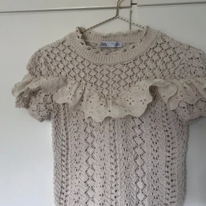 Beige stickad topp med volang från Zara - Supersöt beige stickad topp från Zara med korta ärmar och broderad volangdetalj över bröstet. Mönstrad med hålstickning och rund halsringning. Perfekt för vår och sommar när du vill ha något extra gulligt och unikt.