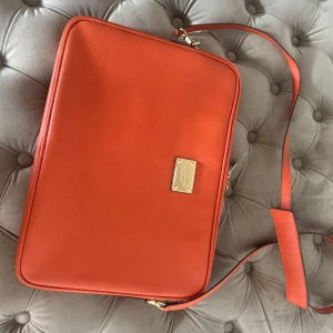Orange datorväska från Michael Kors - Snygg orange datorväska från Michael Kors med guldiga metalldetaljer och logoplatta framtill. Väskan har justerbar axelrem och dubbla dragkedjor med märkets namn. Tillverkad i stilren, rektangulär form. Perfekt för laptop och viktiga grejer.
