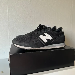 Svarta New Balance sneakers - Klassiska svarta New Balance sneakers med vit logga på sidan och vit detalj på hälen. Skorna har snörning, rund tå och platt sula. Ovandelen är i en mix av mocka och syntetmaterial för en sportig och stilren look.