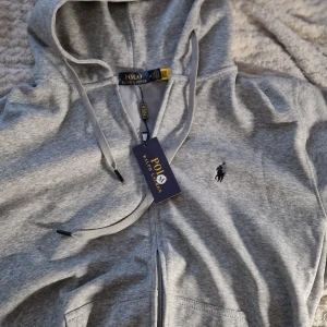 Grå hoodie från Polo Ralph Lauren - (Den är oanvänd och ny, prislappen går med, säljer den eftersom den är för liten på mig och den passar också folk i S bra) Snygg ljusgrå hoodie från Polo Ralph Lauren med klassisk broderad logga på bröstet. Tröjan har huva med snörning.