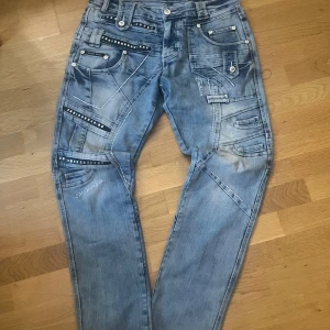 Blå jeans med detaljer och nitar - Säljer ett par ljusblå jeans med coola sömmar, nitar och dekorativa band. Byxorna har flera fickor med knappar och dragkedjor, samt broderad text på ena benet. Snygg streetstil med unik design och raka ben.