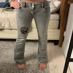 Grå bootcut jeans med slitningar - Grå jeans med bootcut passform och coola slitningar på låret och bakfickan. Jeansen har klassisk femficksdesign, dubbla knappar i midjan och råa detaljer. Perfekta för dig som gillar en edgy och avslappnad stil.
