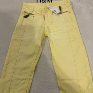 Gula jeansbyxor från H&M   - Snygga ljusgula jeansbyxor från H&M med raka ben och coola dragkedjor vid fickorna framtill. Byxorna har klassisk femficksdesign, bälteshällor och är tillverkade i bomull. Perfekta för dig som vill sticka ut med färg i din outfit.
