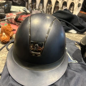 Blå Samshield ridhjälm med metallicdetalj - Snygg svart ridhjälm från Samshield med cool metallicdetalj framtill och stilren design. Hjälmen har en rundad form och bred skärm, perfekt för dig som vill ha både säkerhet och stil på ridbanan. Passar dig som gillar klassisk look med modern touch.
