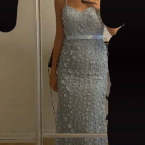 Ljusblå långklänning med blommor - Säljer en ljusblå långklänning med smala axelband och v-ringad urringning. Klänningen har ett vackert blommigt 3D-mönster över hela tyget och ett satinband i midjan.