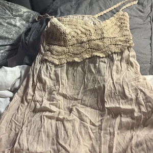 Beige kortklänning med virkad topp - Säljer en söt beige kortklänning med virkad överdel och smala axelband. Kjoldelen är i mjukt, tunt tyg som faller fint och har en lätt skrynklig look. Perfekt för varma dagar och har en bohemisk vibe med sin virkade detalj upptill.