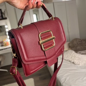 Vinröd axelväska med gulddetaljer - Snygg vinröd axelväska i skinnimitation med justerbar rem och handtag. Väskan är i väldigt bra skick!!💗