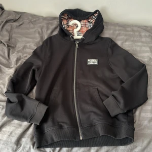 Svart Burberry zip hoodie perfekt för höst och vinter - Säljer en svart hoodie från Burberry i storlek L med klassiskt rutig foder i beige, svart och rött. Tröjan har huva, dragkedja framtill och Burberry-logga på bröstet. Perfekt för dig som vill ha en stilren och lyxig streetwear-look. Hoodien är stor och tjock av sig. Perfekt för att hålla värmen under de kommande årstiderna