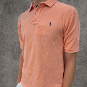 Persikofärgad pikétröja Ralph Lauren S - Säljer en persikofärgad pikétröja från Polo Ralph Lauren i storlek S. Klassisk design med korta ärmar, krage och knappar framtill. Tröjan har den ikoniska blå loggan broderad på bröstet och är gjord i mjuk bomull. Perfekt för en chill och stilren look.