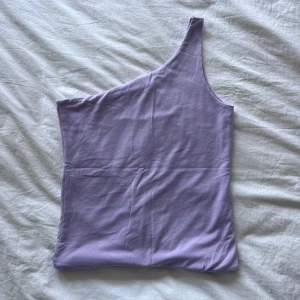 Ljuslila oneshoulder topp body - Säljer en ljuslila body med oneshoulder-design. Toppen är ärmlös och har en clean, slät look utan mönster eller detaljer. Materialet känns mjukt och stretchigt, perfekt för att styla med jeans eller kjol. Färgen är superfräsch och ger en chill vibe.