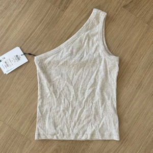 Beige oneshoulder topp från Selected Femme - Snygg ribbad topp i beige melange från Selected Femme. Modellen är oneshoulder med en axel och har en stilren, tight passform. Perfekt för dig som gillar minimalistisk stil och vill ha något som sticker ut lite extra. Materialet är mjukt och stretchigt bomull. Aldrig använt plus prislapp på.