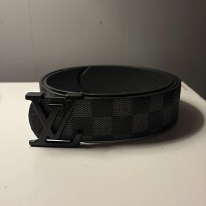 Svart Louis Vuitton rutigt bälte - Snyggt svart bälte från Louis Vuitton med klassiskt rutigt mönster och en stor LV-logga som spänne. Bältet är gjort i skinn och har en stilren, modern vibe som passar till jeans eller kostymbyxor. Perfekt för dig som gillar exklusiva accessoarer.