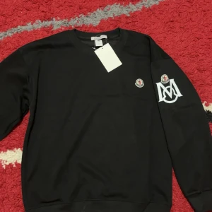 Svart sweatshirt från Moncler - Svart sweatshirt från Moncler med rund hals och lång ärm. Tröjan har Moncler-logga på bröstet och ett stort vitt M-tryck på ena ärmen. Tillverkad i mjuk bomull, perfekt för chill dagar. Klassisk och stilren design med ribbade muddar.