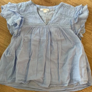 Ljusblå blus med volang från Gina Tricot - Supersöt ljusblå blus från Gina Tricot med volangdetaljer på ärmarna och v-ringad hals. Blusen har broderade partier upptill och är tillverkad i ett mjukt, luftigt bomullstyg. Perfekt för dig som gillar romantisk och chill stil.