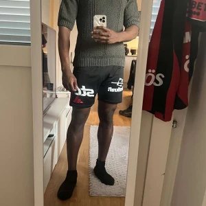 Svarta sportshorts med tryck - Svarta sportiga shorts från Starkraft med vita och röda tryck på framsidan. De har en loose passform och är perfekta för träning eller fotboll. Materialet är lätt och troligtvis polyester, vilket gör dem sköna att röra sig i. Snyggt och stilrent designade för dig som gillar sport.