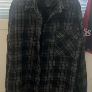 Svart och grå rutig overshirt - Snygg overshirt i svart och grått med rutigt mönster. Skjortan har klassisk krage, knappstängning framtill och en bröstficka med lock. Perfekt att slänga över en t-shirt för en chill look.