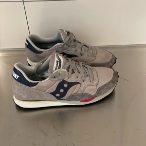 Saucony grå sneakers strl 44 - Saucony sneakers i grått med mörkblå detaljer och vit sula. Skorna har klassisk snörning, rund tå och inslag av mocka på ovansidan. Snygg retrostil med en liten röd detalj vid sulan. Perfekt för dig som gillar sportig och avslappnad look.