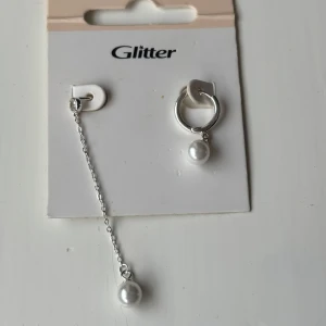 Silverörhängen med pärlor från Glitter - Snygga örhängen i silverfärg från Glitter, ena med en rund creol och hängande vit pärla, andra med lång kedja och pärla längst ner. Perfekt för dig som gillar att mixa och matcha olika längder och detaljer. Endast den runda creol är använd fåtal gånger. 