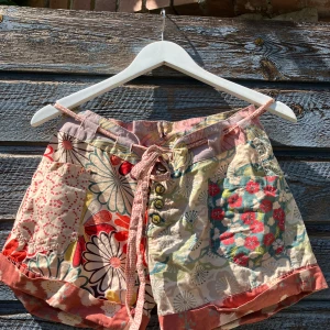 Blommiga shorts från Desigual med snörning - Färgglada shorts från Desigual med blommigt mönster i rött, grönt, beige och rosa. De har snörning i midjan, knappar framtill och flera fickor. Materialet är mjuk bomull och passformen är loose. Perfekta för sommarens chill dagar.