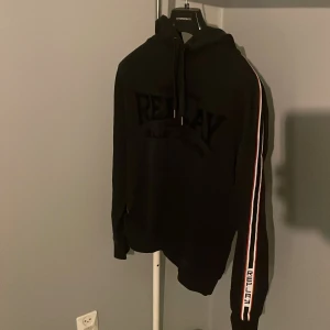 Svart hoodie från Replay med ränder - Svart hoodie från Replay med huva och snörning. Långa ärmar med vita och röda ränder längs sidorna och Replay-logga på ärmslutet. Stor Replay-text på bröstet. Perfekt för chill och streetwear. Mjuk och skön bomullskänsla. Hör av er vid funderingar🤗