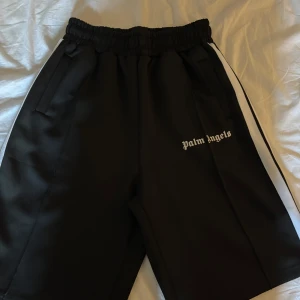 Palm Angels svarta shorts  - Svarta shorts från Palm Angels med vit stripe längs sidorna och logotyp broderad på vänster ben. Modellen har elastisk midja, Materialet känns som polyester. Väldigt bra skick 10/10