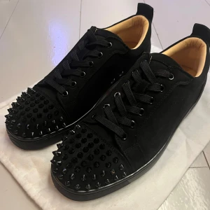 Christian Louboutin svarta sneakers med nitar - Svarta sneakers från Christian Louboutin i mocka med svarta nitar och klassisk röd sula. Skorna har snörning och rund tå, samt en mjuk innersida i beige. Perfekt för dig som vill vara grish.