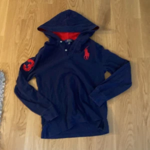 Marinblå hoodie från Polo Ralph Lauren - Snygg marinblå hoodie från Polo Ralph Lauren med röd insida i huvan och rött broderat logomärke på bröstet. Långärmad med röd siffra 3 på ärmen. Mjuk bomullskvalitet och klassisk passform, perfekt för chill dagar.