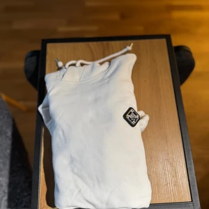 Vit hoodie från J.Lindeberg - Snygg vit hoodie från J.Lindeberg med svart patch på bröstet och klassisk känguruficka. Tröjan har huva med snörning och långa ärmar. Har lite märken samt ett hål i ärmen