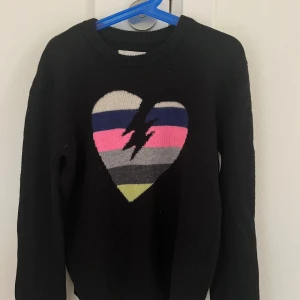 Svart tröja med hjärta från Zadig & Voltaire - Cool svart stickad tröja från Zadig & Voltaire med ett stort hjärta i flera färger och blixt på bröstet. Tröjan har rund hals och långa ärmar. Materialet är mjuk ull och kashmir, perfekt för dig som gillar statement-plagg med färg och edge. 