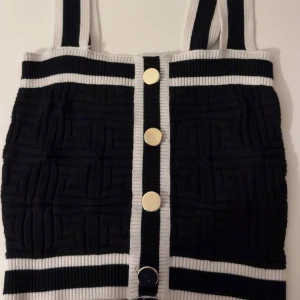 Svart och vit ribbad linnetopp med knappar - Snygg linnetopp i svart och vitt med breda axelband och ribbade detaljer. Toppen har ett coolt geometriskt mönster och stora guldfärgade knappar framtill. 