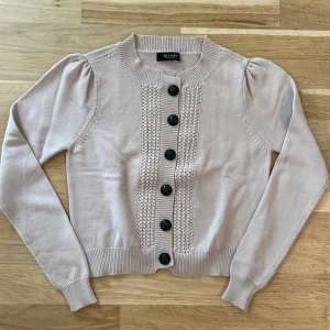 Beige kofta från Sand, M - Snygg beige kofta i lite kortare modell från Sand i storlek M. Koftan har puffade axlar, dekorativa svarta knappar och ett stickat mönster framtill. Materialet är mjukt med 79% polyamid och 21% viskos. 