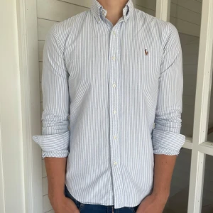Randig skjorta från Ralph Lauren - Säljer denna riktigt snygga vit och blå randiga Ralph lauren skjorta, den är i mycket bra skick förutom att armhålorna har lite deofläckar annars mycket bra skick, storlek S modell slim fit, den är 72 cm lång och 50 cm bred under armhålorna, hör gärna av dig vid frågor!