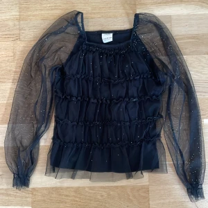 Svart glittrig blus från Zara stl 152 - Supersnygg svart blus från Zara med transparenta långa ärmar och glittriga detaljer. Toppen har flera lager med volanger och ett mesh-tyg som ger en cool och festlig vibe. Bara använd nån enstaka gång.