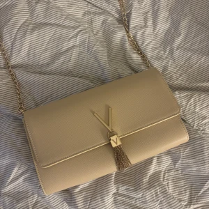 Beige axelväska från Mario Valentino - Snygg beige axelväska från Mario Valentino med guldfärgad kedja och V-detalj framtill. Har använt den en gång till balen och kommer inte mer till användning, den har inte några fläckar eller sånt