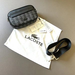 Svart axelväska från Lacoste - Snygg svart axelväska från Lacoste med geometriskt mönster i grått och silver. Väskan har justerbar axelrem, dragkedja med Lacoste-logga och krokodilemblem på framsidan. Materialet är skinn och polyester, perfekt för dig som gillar stilrena detaljer.