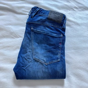 Replay Anbass blå jeans, storlek 31 - Snygga Replay Anbass jeans i klassisk blå tvätt med diskreta slitningar. Modellen har fem fickor, normal passform och raka ben. Jeansen är tillverkade i mjukt denim-material med dragkedja och knapp i midjan. Perfekta för dig som gillar stilrena och tidlösa jeans. Midjemått: 40 cm benlängd: 101 cm !! Priset kan diskuteras !!