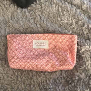 Rosa mönstrad plånbok Louise.C - Snygg plånbok från Louise.C i rosa och vitt med grafiskt mönster. Perfekt storlek för smink, småprylar eller pengar. Stängs med dragkedja upptill och har en mjuk, flexibel form. Märke syns tydligt på framsidan.